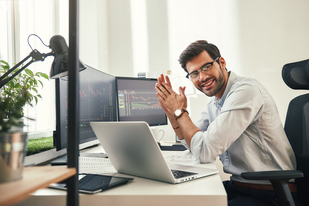 smiling man with glasses sits at desk in front of monitors and his laptop