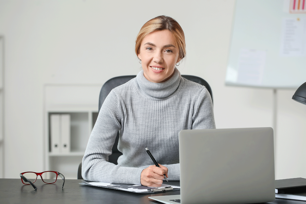 young woman smiling as she sits at a laptop and contemplates what to look for in an EMR software system for behavioral healthcare.