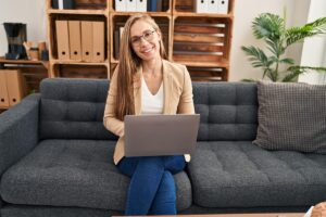 a smiling behavioral healtcare worker is seated on a couch while working on a laptop to understand the impact of regulatory shifts on behavioral health billing