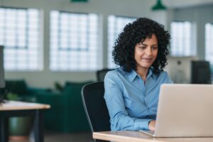 Professional woman sitting at a desk in a modern office, focused on working on her laptop