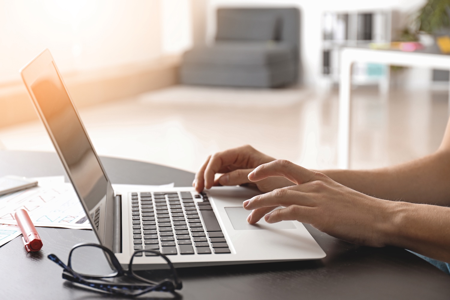 Person typing on a laptop at a table with glasses and papers nearby, working in a bright home or office space