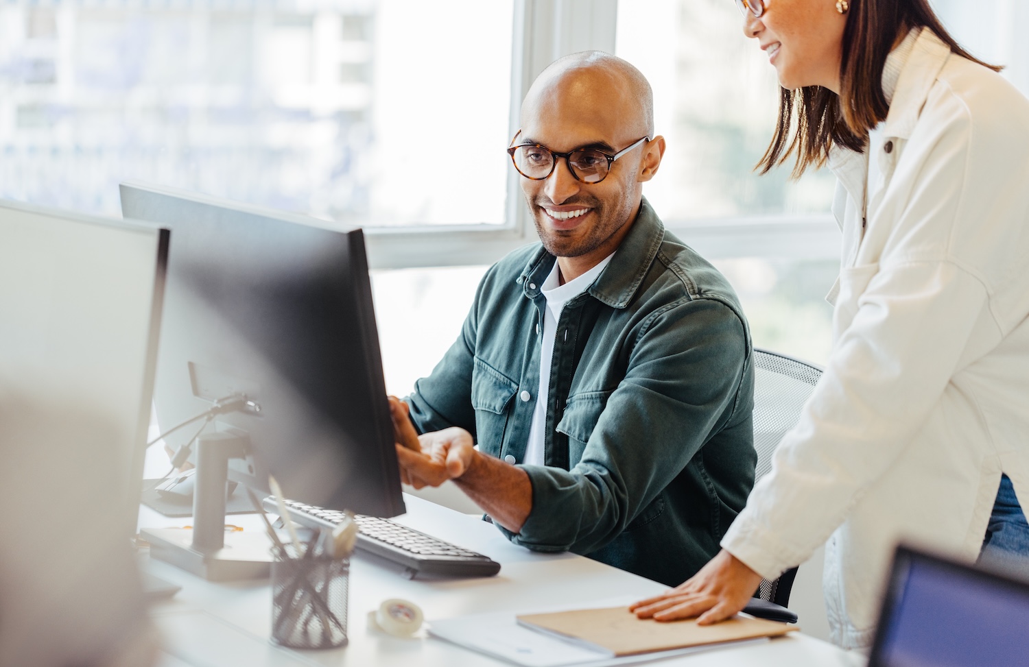 Two colleagues collaborating at a desk, one sitting at a computer and smiling while the other stands beside him, discussing work